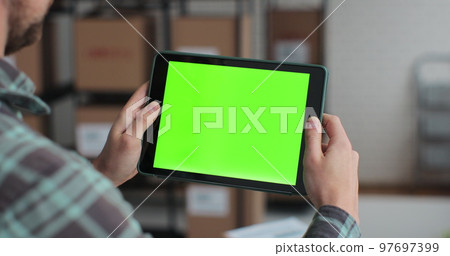 Man using tablet computer with green screen chromakey. Browsing the Internet in a warehouse full of cardboard boxes. 97697399