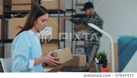 A female support service manager checks the order number on the parcel using a computer, prepares a box for mailing. Warehouse work. 97697435