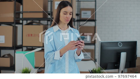 Portrait of a woman using a smartphone at work in a warehouse. A woman connects to customer support via the Internet. Work manager in an online store. Portrait of a woman using a smartphone at work in a warehouse. A woman connects to customer support via the Internet. Work manager in an online store. 97697464