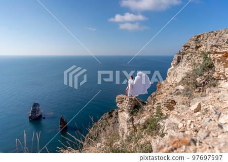 A beautiful young woman in a white light dress with long legs stands on the edge of a cliff above the sea waving a white long dress, against the background of the blue sky and the sea. 97697597