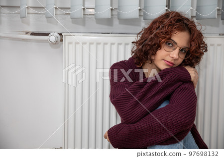 The girl, worried about the constant lack of heating, sits by the heater. The girl, worried about the constant lack of heating, sits by the heater. 97698132