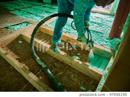 Male builder insulating wooden frame house. Cropped view of man worker spraying polyurethane foam on floor inside of future cottage, using plural component gun. Construction and insulation concept. Male builder insulating wooden frame house. Cropped view of man worker spraying polyurethane foam on floor inside of future cottage, using plural component gun. Construction and insulation concept. 97698190