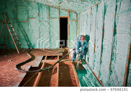 Male builder insulating wooden frame house. Man worker spraying polyurethane foam inside of future cottage, using plural component gun. Construction and insulation concept. 97698191