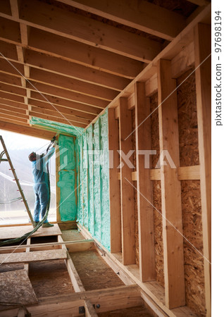 Male builder insulating wooden frame house. Man worker spraying polyurethane foam inside of future cottage, using plural component gun. Construction and insulation concept. Male builder insulating wooden frame house. Man worker spraying polyurethane foam inside of future cottage, using plural component gun. Construction and insulation concept. 97698194