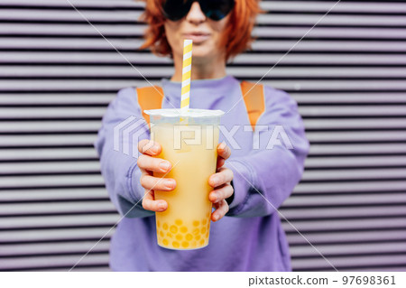 Glass of fruity sugar flavored tapioca pearl bubble tea with straw in hand of hipster fashion woman in sunglasses and purple clothes on gray striped wall background. Selective focus. Copy space 97698361