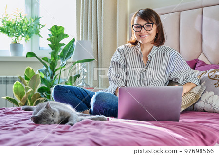 Middle aged woman at home on bed with laptop and cat Middle aged woman at home on bed with laptop and cat 97698566