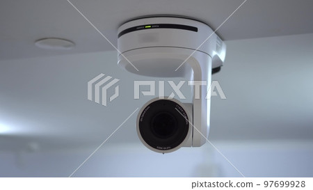 The remote controlled camera uses an electromechanical optical zoom. A white camera hangs from the ceiling in the room. Video camera for streaming and video recording. The remote controlled camera uses an electromechanical optical zoom. A white camera hangs from the ceiling in the room. Video camera for streaming and video recording. 97699928