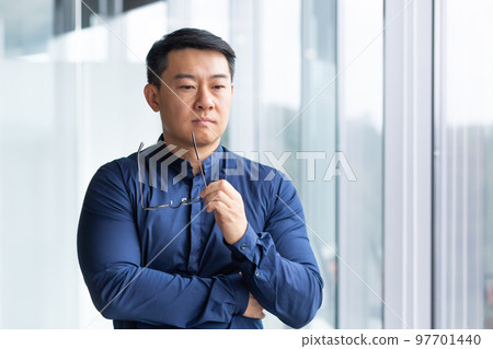 Portrait of a serious young Asian businessman, lawyer. He stands thoughtfully in the office by the window, holds glasses in his hands, looks to the side. Portrait of a serious young Asian businessman, lawyer. He stands thoughtfully in the office by the window, holds glasses in his hands, looks to the side. 97701440