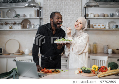 Cheerful married multinational couple using laptop while cooking healthy food in kitchen 97702609