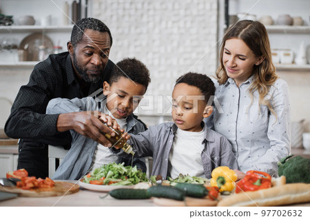 Happy multiracial parents and their small boys kids have fun preparing healthy food at home. 97702632