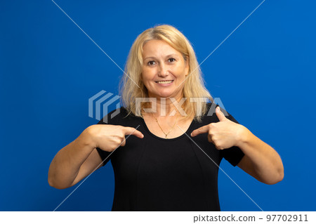 Portrait of a charming happy woman in a black t-shirt smiling and pointing at herself, proud of her own success, bragging about her achievements. Indoor studio shot isolated on blue background 97702911