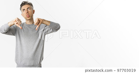 Image of skeptical and disappointed middle-aged man, showing thumbs-down and sticking tongue, judging something very bad and lame, standing over white background 97703019