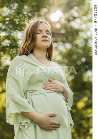 Young pregnant female holds hands on big belly, closed eyes, green trees,sky, sunlight on background. Happy female wears dress. Magic pregnancy. Labor, emotional connection with baby. Vertical 97703104
