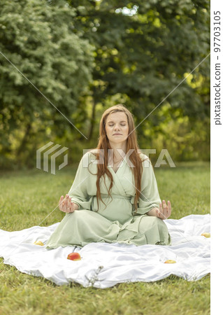 White pretty pregnant woman meditates in park, sits on blanket, stress relief,mindfulness. Enjoying meadow,relaxation. Healthy lifestyle. Labor, selfcare. Fresh air,green trees on background. Vertical 97703105