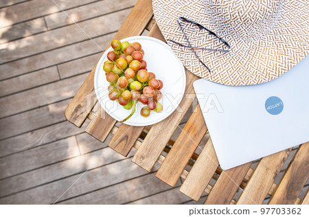 Closed laptop, grape, stylish hat, sunglasses are on a wooden table on a summer terrace with copy space. Summer mood, vacation, freelance, distance work concept. Top view. Background with copy space. 97703362