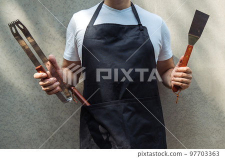 A man wearing black chef's apron, holding barbecue tools 97703363
