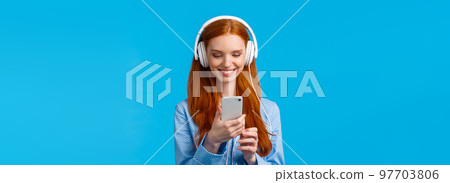 Technology addication, lifestyle and women concept. Cheerful good-looking redhead woman in nightwear, wearing big headphones listen music, smiling pick songs in smartphone app 97703806