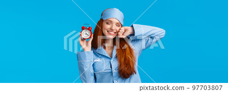 Girl slept well, wake up energized, sleepy stretching with closed eyes and lovely smile, holding red cute clock, set up alarm to be work in time, wearing pyjama and sleep mask, blue background 97703807