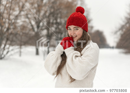 Sly smiling girl in red knitted hat and mittens in snowy winter park. Portrait of young woman on walking 97703808