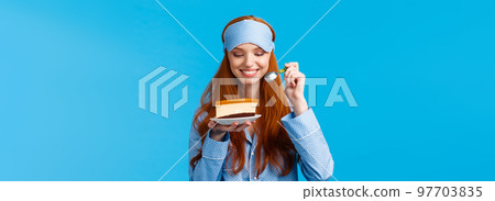 Waist-up portrait cheerful and delighted young caucasian redhead woman in pyjama and sleep mask, holding slice cake and spoon smiling delighted, looking at tasty dessert, wear nightwear 97703835