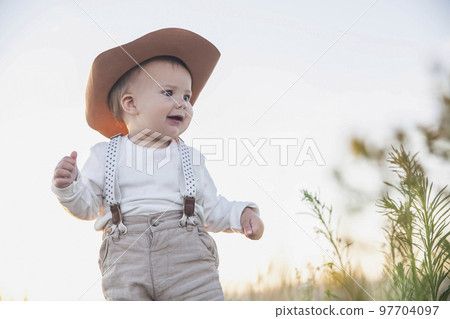 baby in a hat and suspenders looking in the field at sunset 97704097