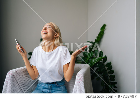 Front view of happy amazed young woman holding smartphone feeling excited winning online lottery bid on cellphone sits on armchair at home. Excited female reading online mobile phone content at home. Front view of happy amazed young woman holding smartphone feeling excited winning online lottery bid on cellphone sits on armchair at home. Excited female reading online mobile phone content at home. 97718497