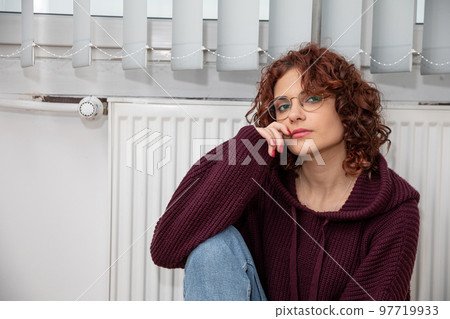 The girl, worried about the constant lack of heating, sits by the heater. The girl, worried about the constant lack of heating, sits by the heater. 97719933