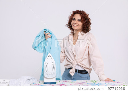 The girl smiles gratefully while standing by the ironing board and iron. The girl smiles gratefully while standing by the ironing board and iron. 97719938