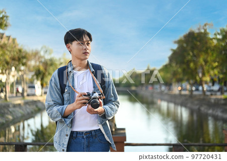 Young asian male blogger with digital camera standing on bridge with scenery view of river and beautiful blue sky 97720431