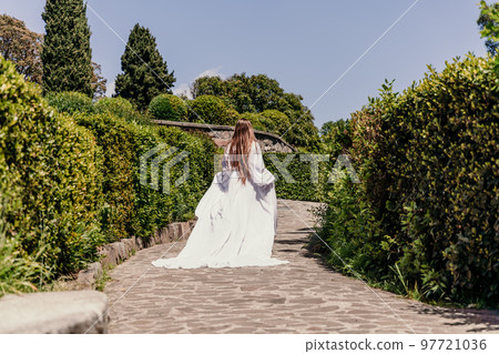 a beautiful woman with long brown hair and long white dress runs along a path along beautiful bushes in the park rear view a beautiful woman with long brown hair and long white dress runs along a path along beautiful bushes in the park rear view 97721036