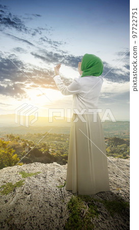 The rear view of a Muslim woman in a veil raising a hand and praying The rear view of a Muslim woman in a veil raising a hand and praying 97722751