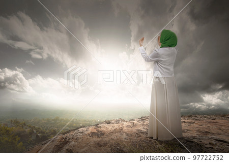 The rear view of a Muslim woman in a veil raising a hand and praying The rear view of a Muslim woman in a veil raising a hand and praying 97722752