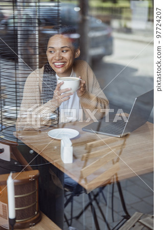 Enthusiastic happy African woman works on laptop, smiles joyfully, contemplates outside the window, enjoys cappuccino 97724207