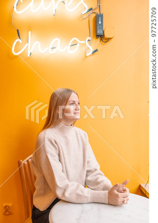 A young woman sits at a table in a cafe and looks out the window while waiting for her coffee or breakfast order A young woman sits at a table in a cafe and looks out the window while waiting for her coffee or breakfast order 97725709