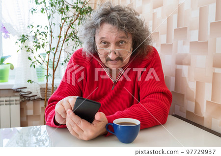 An elderly man in glasses and a phone in his hand is sitting at the table with a cup of coffee 97729937