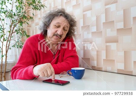 9. A pensioner in a red jumper with a phone in his hands sitting at the table 97729938