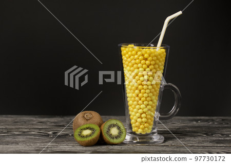 The glass goblet is filled with vitamins in the form of dragees. Kiwi halves nearby. On black pine boards. On a black background. 97730172