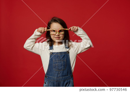 Adorable Caucasian baby girl wearing white pull and blue denim overalls, holding Christmas red and green striped lollipops candy canes like glasses, looking at camera, isolated on colored background Adorable Caucasian baby girl wearing white pull and blue denim overalls, holding Christmas red and green striped lollipops candy canes like glasses, looking at camera, isolated on colored background 97730546