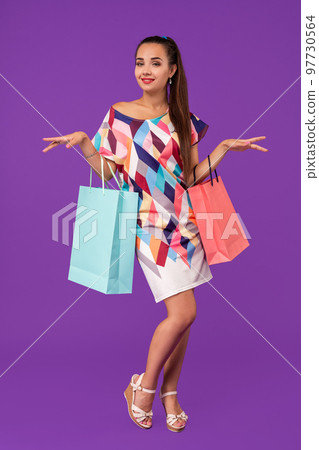 Beautiful young woman holding shopping bags and looking at the camera while standing against purple background. 97730564