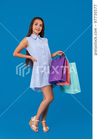 Beautiful young woman holding shopping bags and looking at the camera while standing against blue background. 97730577