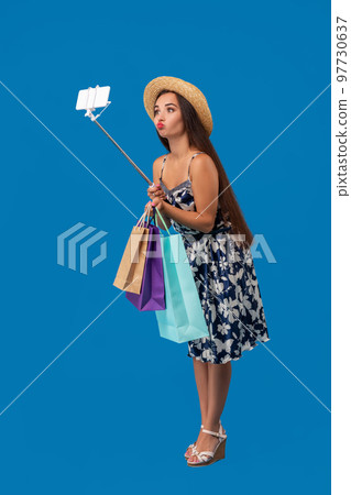 Young woman in casual clothes with shopping bags using selfie stick to take a self portrait on blue studio background with copy space 97730637