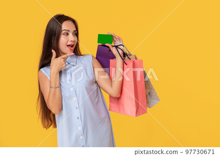 Charming pretty attractive sexy girl showing credit card and holding many colorful packages in hand, it is comfortable to use bank card for shopping, standing over yellow background 97730671