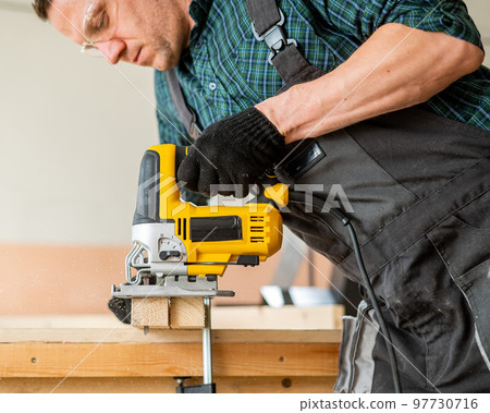 A man cuts a wooden plank with an electric jigsaw in a workshop. A man cuts a wooden plank with an electric jigsaw in a workshop. 97730716