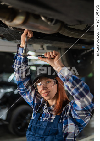 Female mechanic unscrew the nuts on the bottom of the car that is on the lift. A girl at a man's work. 97730735