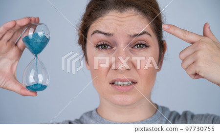 Unhappy caucasian red-haired woman looks at the hourglass and points to the wrinkles on her forehead. Aging concept. 97730755