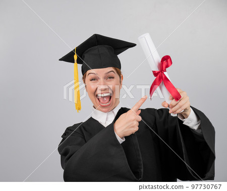 Happy woman in graduate gown pointing finger at received diploma.  97730767