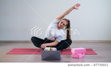Young overweight caucasian woman goes in for sports online by laptop. The girl sits on a mat and does side bends.  97730931
