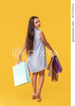 Full length photo of young woman in dress looking back on copy space and holding colorful shopping packages over yellow background Full length photo of young woman in dress looking back on copy space and holding colorful shopping packages over yellow background 97731015