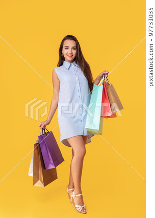 Image of a young brunette lady in white and blue summer striped dress posing with shopping bags and looking at camera over yellow background. Image of a young brunette lady in white and blue summer striped dress posing with shopping bags and looking at camera over yellow background. 97731035