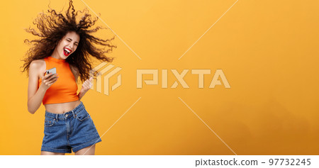 Portrait of joiyful carefree and happy charming european woman with curly hairstyle laughing out loud holding smartphone wearing wireless earbuds listening awesome music via app over orange wall Portrait of joiyful carefree and happy charming european woman with curly hairstyle laughing out loud holding smartphone wearing wireless earbuds listening awesome music via app over orange wall 97732245